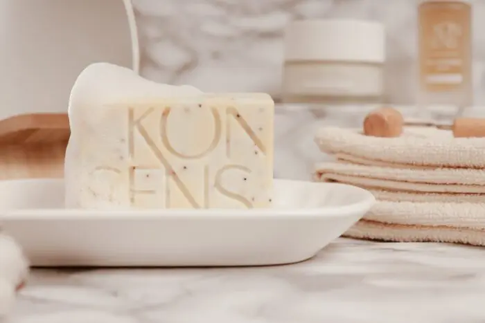 KONSENS Anti-Akne Seife mit feinem Schaum auf einer weißen Seifenschale, im Hintergrund sind Handtücher und Hautpflegeprodukte zu sehen.