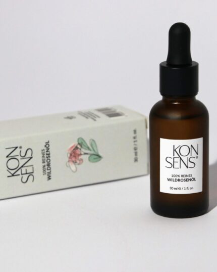 “Flasche mit 100% reinem Wildrosenöl von Konsens, präsentiert neben der eleganten Verpackung, die auf einer Seite liegt. Erhältlich bei Mel’s Beauty & Academy.”